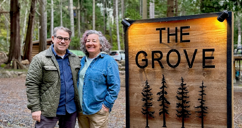 Marc and Dana stand beside a large wooden sign that reads “THE GROVE 684” with tree silhouettes, set in a wooded outdoor area on Mayne Island